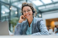 Smiling young businesswoman with curly hair and glasses wearing gray blazer during video call in modern office environment.