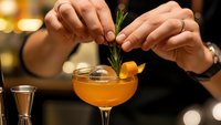 Professional bartender garnishing orange cocktail with fresh herb sprig in elegant coupe glass at upscale bar with warm ambient lighting for hospitality industry.
