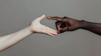 Close up of light and dark-skinned hands reaching towards each other against neutral gray background, symbolizing unity, connection and breaking racial barriers.