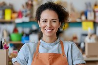 Mixed race female artisan wearing orange apron and denim shirt smiling at camera in creative workshop studio with art supplies and crafting materials in background.