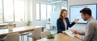 Professional businesswoman presenting financial data to male colleague in modern office meeting room with charts and documents on table.