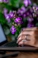 Female hand typing on laptop keyboard with purple flowers in soft focus background creating peaceful home office atmosphere for remote work setting.