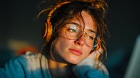 Young woman with glasses and messy hair working late at computer in warm ambient lighting, tired freelancer or student studying at night.