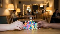 Diverse group of colorful paper figures held by two hands together on wooden table with warm holiday lights and decorated interior background, representing unity, teamwork, community support and togetherness.
