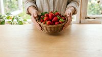 Farmer holding wicker basket filled with fresh ripe strawberries on wooden table, natural daylight from window, organic produce harvest.