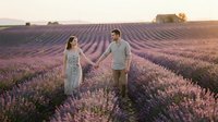 Young couple holding hands in blooming lavender field at golden hour, romantic outdoor engagement or anniversary portrait session in rural countryside landscape.