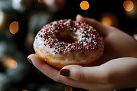 Hands holding glazed donut with festive red and green sprinkles against warm bokeh lights background for holiday celebrations and seasonal treats.