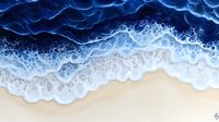 Aerial beach view with deep blue ocean waves crashing on sandy shore, creating white foam patterns and natural swirling textures. Dynamic water meets pristine sand coastline.