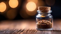 Small glass jar with cork lid filled with ground coffee or spices on wooden surface against blurred warm bokeh lights background creating cozy atmosphere.