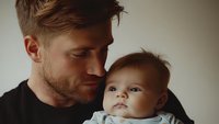 Caucasian father holding infant baby in tender moment against neutral background, showing emotional connection and paternal love in moody lighting.