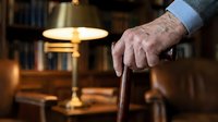 Senior man with aged, wrinkled hands holding wooden cane in warm home library setting with brass lamp and bookshelves, representing aging, mobility, retirement.