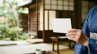 Senior man in denim shirt holding blank white card indoors near traditional wooden architecture and garden view, suitable for messaging and communication concepts.