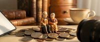 Elderly couple figurines sitting on pile of coins representing retirement savings and financial planning for senior years with warm home office background.