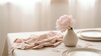 Pale pink peony in white ceramic vase on dining table with place settings and draped fabric for elegant home decor and event styling.