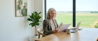 Senior businesswoman reading documents at modern office desk with laptop and plant near large window overlooking countryside fields.