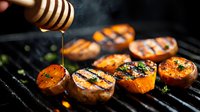Grilled sweet potatoes drizzled with honey on barbecue grill with fresh herbs. Golden honey drips from wooden dipper onto charred orange rounds creating steam in dark setting.
