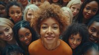 Diverse group of young women smiling at camera, focus on woman with natural curly hair in orange sweater surrounded by multicultural female friends in casual setting.