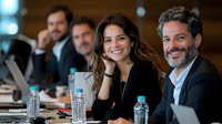Business team at corporate meeting with diverse professionals smiling. Young Hispanic woman and mature executive in foreground, colleagues discussing strategy.