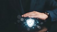 Businessman hands protecting glowing shield icon with digital particles on dark background representing cybersecurity and data protection concepts.