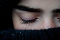 Close-up view of closed eye with long eyelashes and natural freckles, partially covered by dark blue knitted fabric, creating intimate emotional portrait.