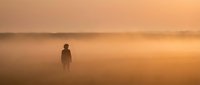 Solitary figure standing in misty field during golden hour sunrise, creating atmospheric silhouette against warm orange sky for contemplative mood concepts.