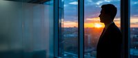 Silhouette of businessman standing by office window at sunset overlooking city skyline, contemplating future success and corporate leadership opportunities.