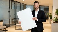 Young Caucasian businessman in black suit holding blank white poster in modern office lobby with glass walls and contemporary furniture for presentation.