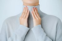 Young Asian woman in light gray turtleneck sweater touching her throat with both hands, showing signs of thyroid problems or sore throat discomfort.