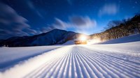 Groomed ski slope with corduroy pattern under starry night sky and mountain backdrop. Winter sports resort scene with snow machine creating powder snow.