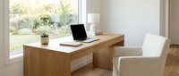 Wooden desk with laptop and potted plant in bright, minimalist home office space with large windows overlooking garden and comfortable chair for remote work.