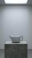 Minimalist concrete teapot on a stone pedestal with TEA inscription, displayed in a modern gallery with soft overhead lighting.