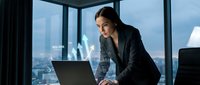Professional businesswoman working on laptop in modern office with city skyline view. Corporate executive analyzing data in contemporary workplace.