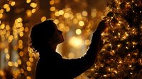Silhouette of woman decorating Christmas tree with golden bokeh lights creating warm festive atmosphere in dark room.