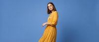 Young Caucasian woman in flowing yellow dress posing gracefully against vibrant blue studio background for fashion and lifestyle photography.