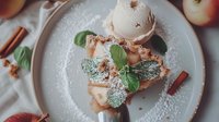 Sweet apple pie slice topped with vanilla ice cream, fresh mint leaves and powdered sugar on white plate, decorated with cinnamon sticks, overhead view.
