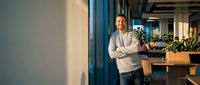 Confident middle-aged Caucasian businessman with crossed arms standing in modern office space with blue pillars and plants in background.