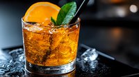 Refreshing cocktail with orange slice and mint garnish in rocks glass on dark background with ice cubes, golden drink glowing in moody lighting.