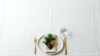 Elegant table setting with gold cutlery, white plates, fresh greenery and dried citrus slice on linen tablecloth for fine dining and event planning.