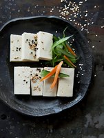 Fresh tofu cubes garnished with sesame seeds, sliced carrots and green onions served on black ceramic plate against dark rustic background. Asian cuisine.