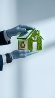 Hands in protective gloves holding miniature eco-friendly house model with solar panels, moss covering and recycling symbol for sustainable living concepts.