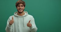 Young man in white hoodie showing thumbs up against green background, expressing positivity and confidence with genuine smile. Great for lifestyle and advertising.