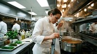 Female chef in white uniform tasting steaming dish in busy professional restaurant kitchen, culinary expertise and passion on display.