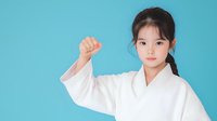 Young Asian girl in white karate uniform showing fist gesture against turquoise background, representing martial arts training and confidence.