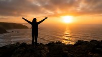 Woman with arms raised celebrating freedom and success at sunset on rocky coastline overlooking ocean. Concept for motivation, achievement, wellness.