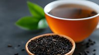 Aromatic black tea leaves in ceramic bowl and freshly brewed tea in orange cup on dark background with green tea leaves, selective focus composition.