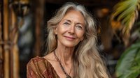 Senior Caucasian woman with long gray hair smiling gently, wearing earth-toned clothing against tropical background. Natural aging beauty portrait.