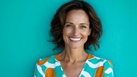 Cheerful middle-aged Caucasian woman with short wavy brown hair wearing colorful patterned dress smiling confidently against turquoise background.
