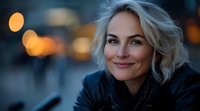 Smiling blonde woman with natural makeup outdoors at dusk, warm bokeh lights in background creating atmospheric evening portrait with shallow depth of field.