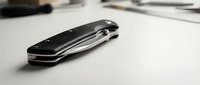 Black carbon fiber folding pocket knife with steel blade closed on white surface. Modern tactical utility tool for outdoor activities and everyday carry.