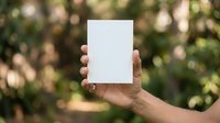 Caucasian male hand holding blank white card mockup outdoors with blurred green foliage background for branding and marketing purposes.